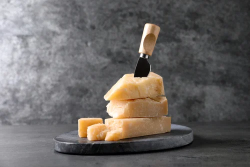 Na imagem há um pedaço de queijo grana padano com uma faca e lascas de grana padano ao lado