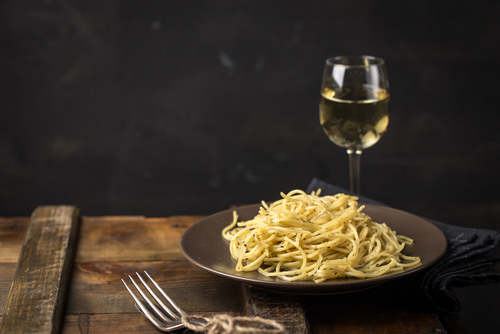 Na imagem, há um prato com macarrão espaguete à carbonara ao lado de uma taça de vinho branco