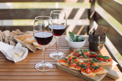 Na imagem, há uma mesa de jardim com aperitivos e duas taças de vinho tinto